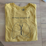 Yellow t-shirt with a graphic of lungs and text 'so many things to do' on a wooden surface.